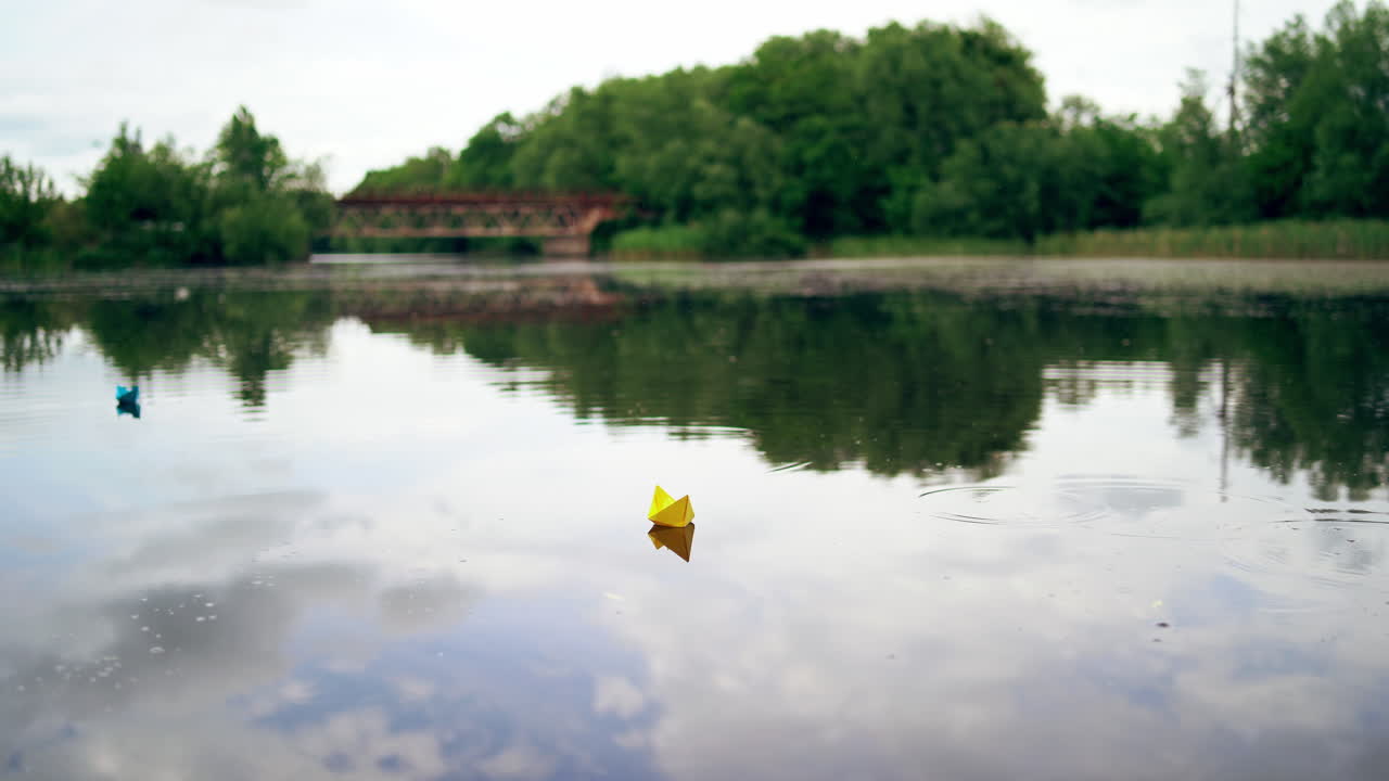 Handmade yellow and blue paper ships are floating on the river in the evening. Two origami boats are sailing on calm river with a little rain falling from the sky into the water.