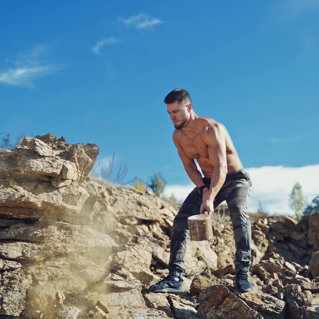 Strong man is training with heavy hammer. Athletic sportsman breaking stones with iron hammer on the natural rocky background. Slow motion.