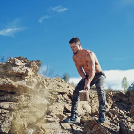 strong man is training with heavy hammer. athletic sportsman breaking stones with iron hammer on the natural rocky background. slow motion.