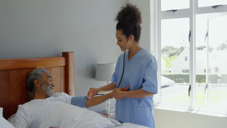 Front-view-of-young-mixed-race-female-doctor-checking-blood-pressure-of-senior-man-on-bed-at-home-4k