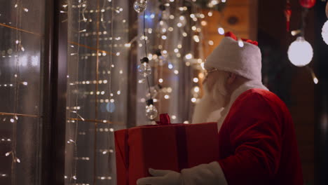 a real santa claus with glasses and a beard on the outdoor comes to the window of the house with a gift on the night before christmas