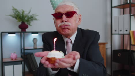 senior businessman celebrating lonely birthday in office, blowing candle on small cake making a wish