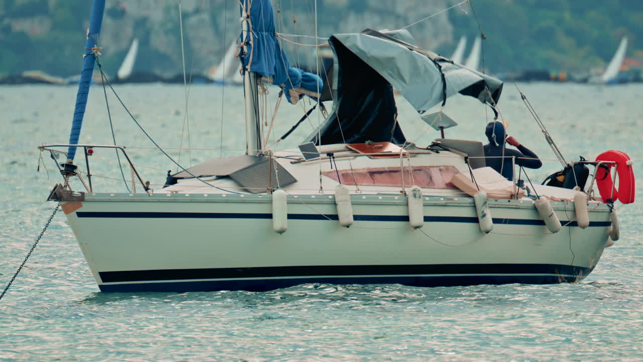 View of a small sailboat rocking on the waves with a lone sailor on deck, diver preparing to jump into sea