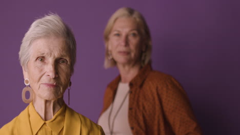 blonde senior woman with short hair wearing mustard colored shirt and jacket and earrings, posing with blurred mature woman on purple background