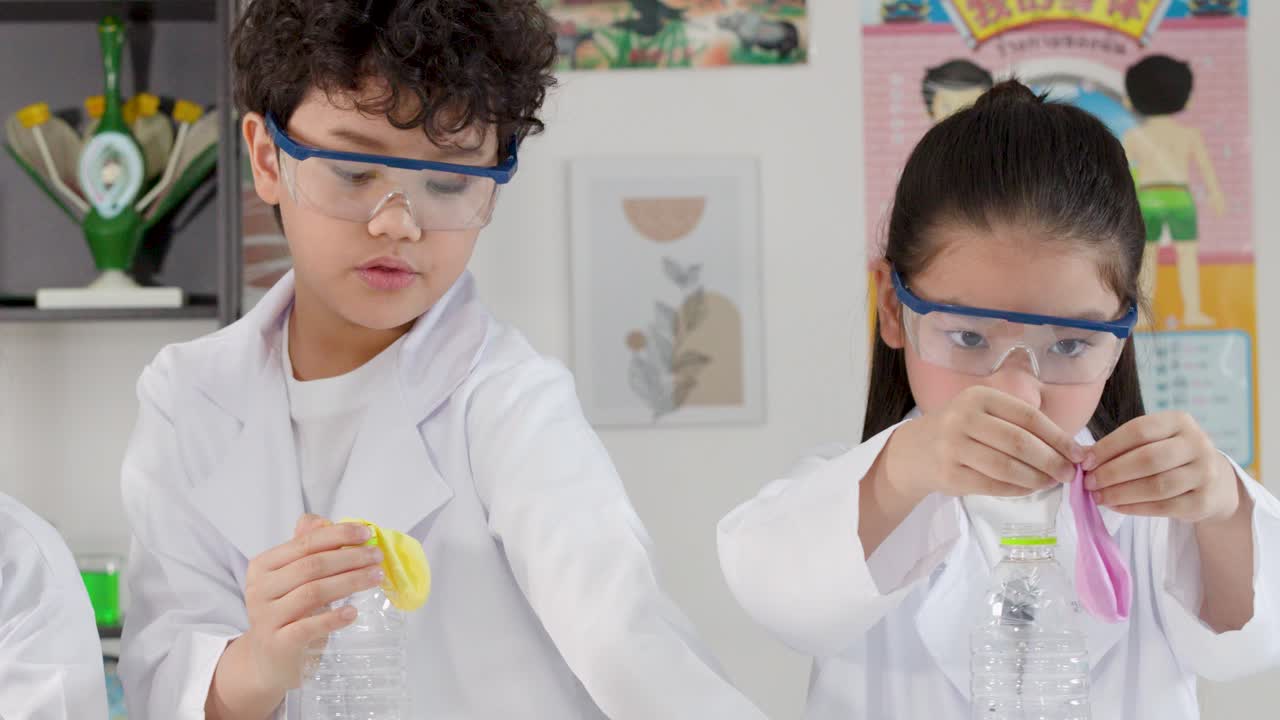 Two young Asian students wear safety goggles while attaching balloons to plastic bottles in classroom