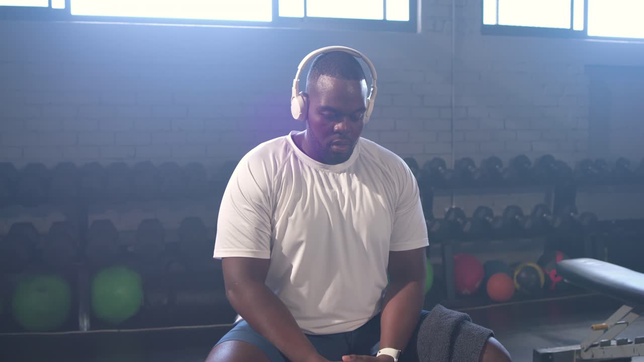 Pria Afrika-Amerika memeriksa jam tangan, memakai headphone di bangku di gym, bersiap untuk latihan.