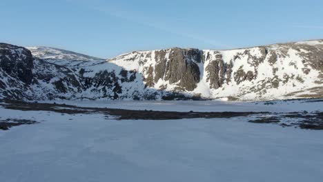 Imágenes-De-Drones-Aéreos-Que-Se-Elevan-Sobre-El-Lago-Congelado-Etchachan-En-Invierno-Y-Vuelan-Hacia-Montañas-Cubiertas-De-Nieve-Y-Un-Acantilado-Vertical-De-Roca-Escarpada,-Nieve-Y-Una-Cresta-Helada