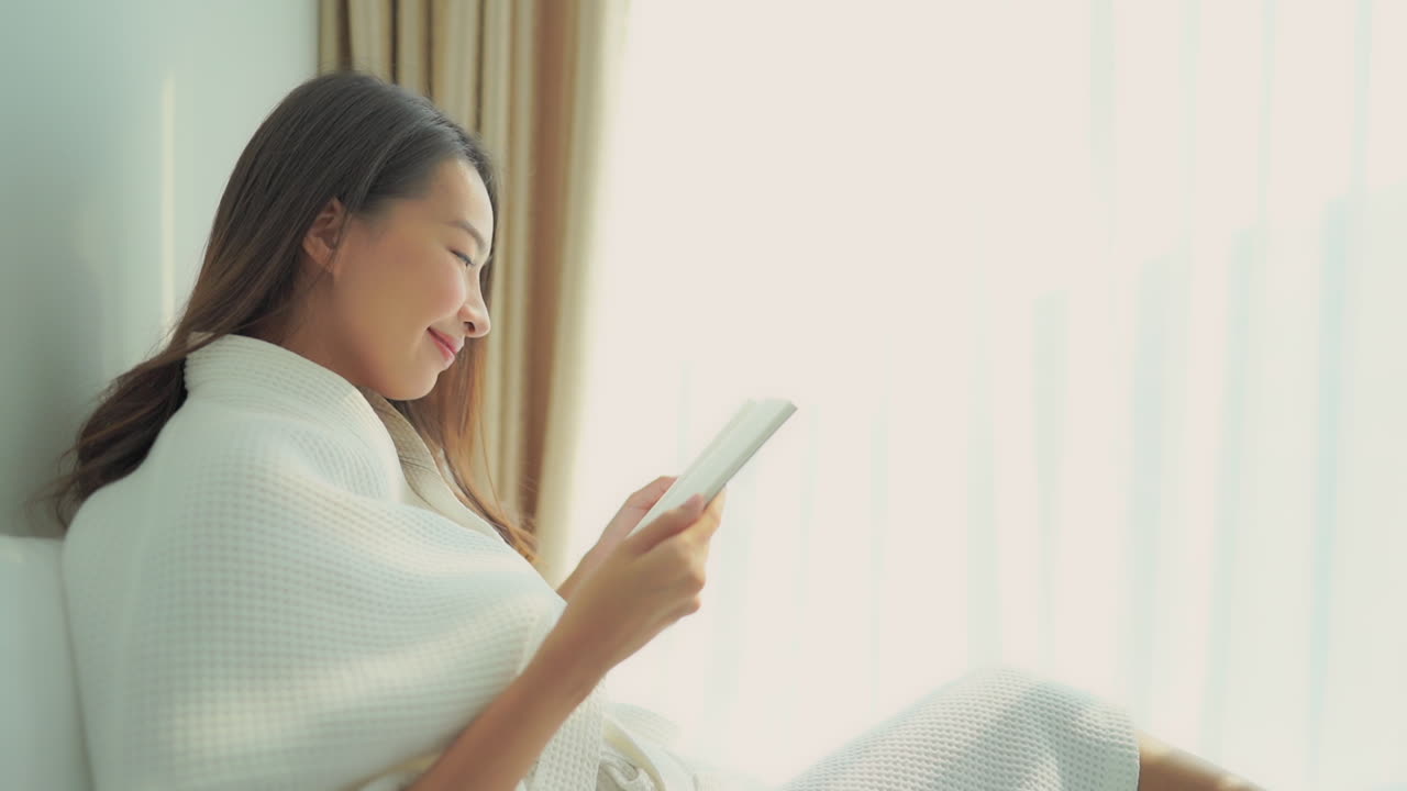 Close up of women wrapped up in a resort robe uses the light from the window to read
