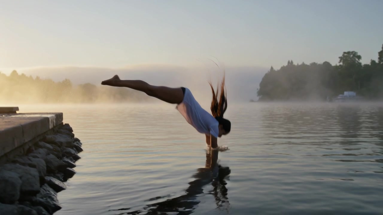 Donna in maglietta bianca e pantaloncini scuri si sporge da un molo di legno all'alba, tuffandosi nel lago.