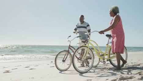 happy senior african american couple walking with bikes at beach, in slow motion