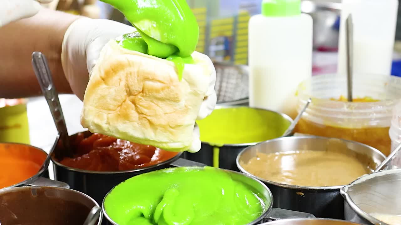 Primo piano di un venditore che applica una vivace crema pasticcera verde sul pane tra varie guarnizioni colorate.