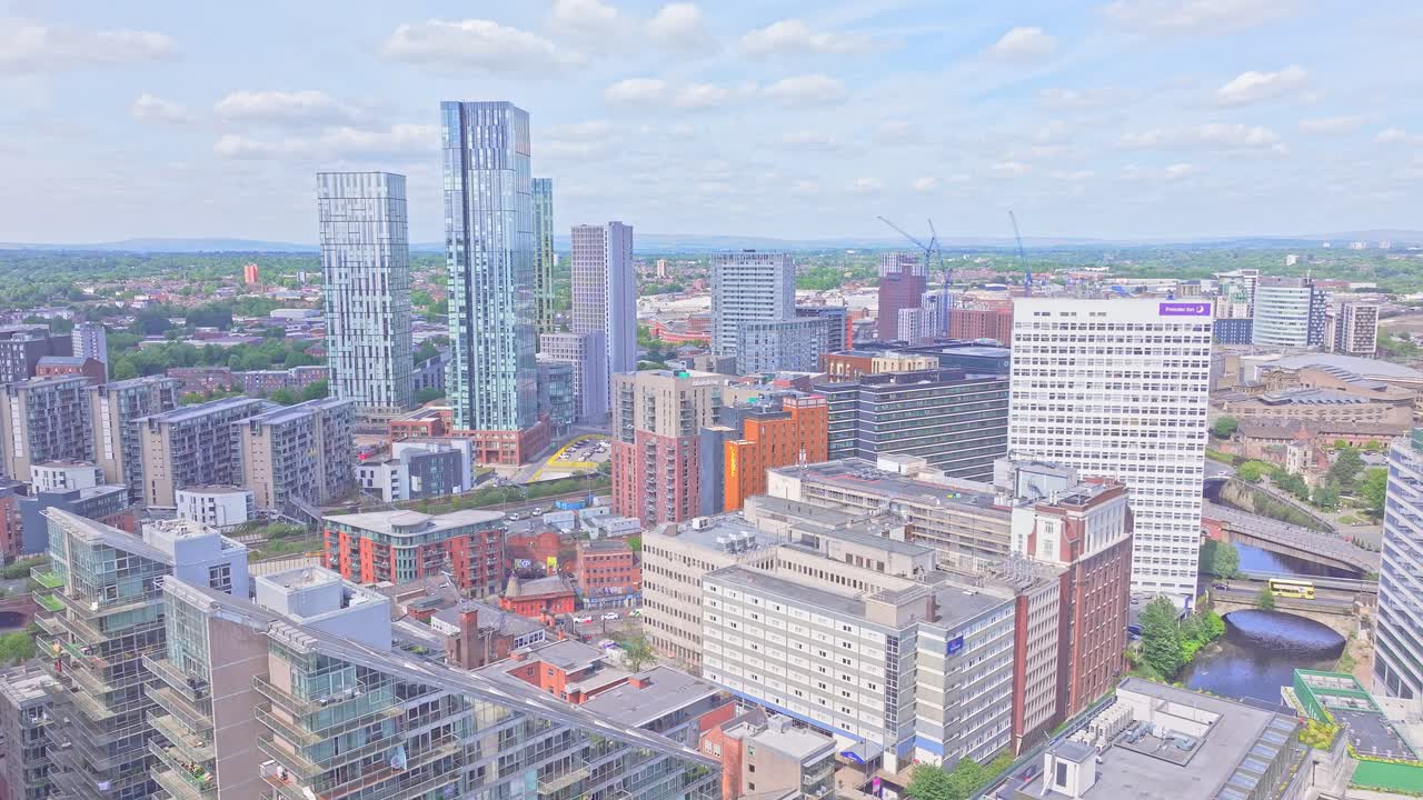 Aerial drone shot slowly ascending above Manchester city centre showcasing Beetham Tower, City Tower, Deansgate Square, and the Premier Inn building under a vibrant partly cloudy sky