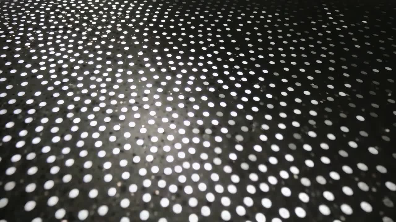 Spostamento della camera durante le riprese di un pannello forato e macchiato in primo piano, rivelando puntini luminosi che si illuminano e poi si attenuano
