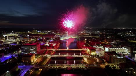 Downtown-Des-Moines,-Iowa-buildings-at-night-with-fireworks-exploding-in-celebration-of-Independence-Day-over-bridges-with-drone-video-stable