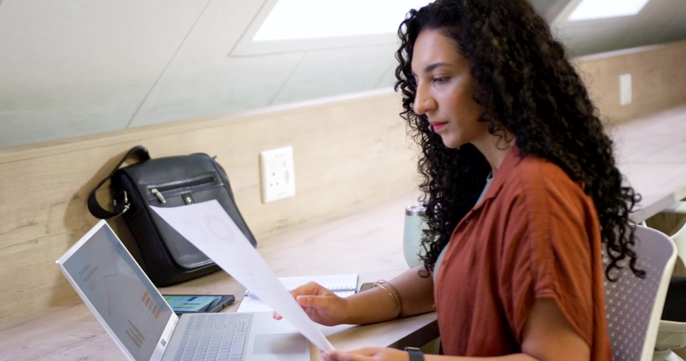 Donna afroamericana che digita e confronta grafici stampati con un laptop a una scrivania di legno per un rapporto.