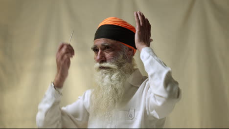 Fotografía-De-Estudio-De-Un-Hombre-Sikh-Mayor-Con-Barba-Que-Usa-Una-Aguja-De-Salai-Mientras-Se-Pone-Un-Turbante-Contra-Un-Fondo-Liso,-Filmada-En-Tiempo-Real