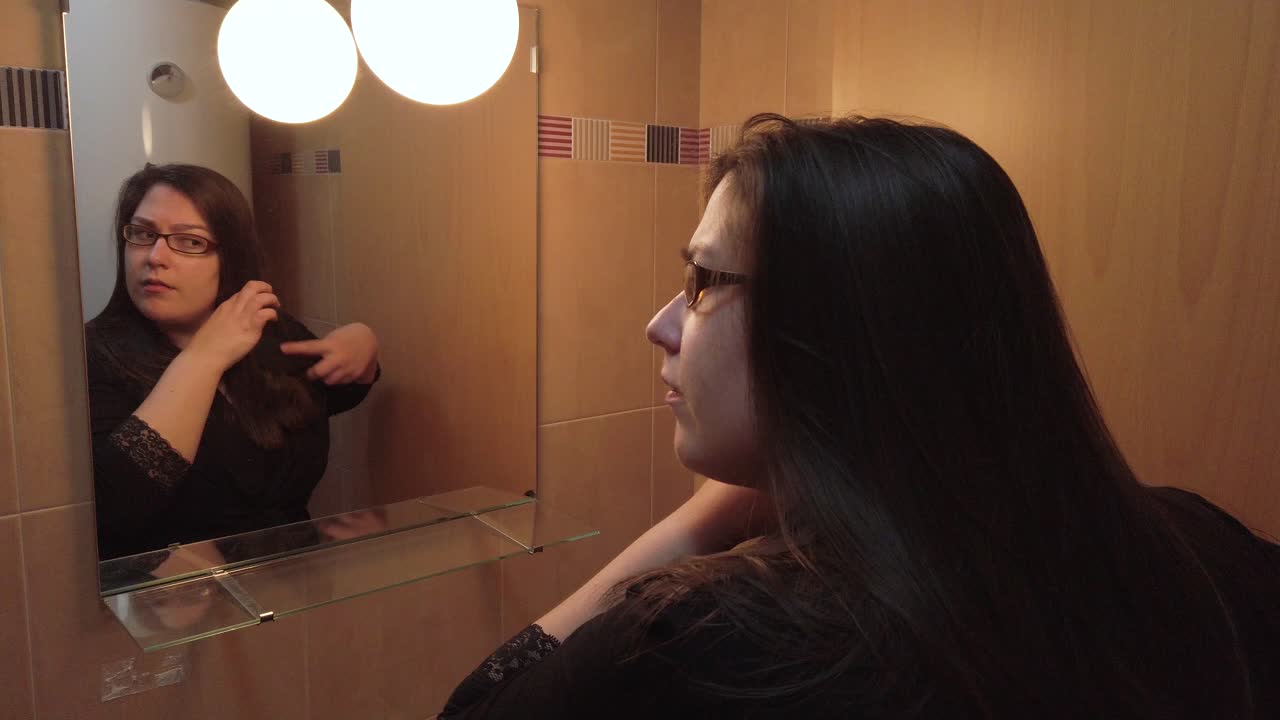 Donna che si spazzola i capelli lisci e castani nello specchio del bagno, vista da dietro con riflesso.