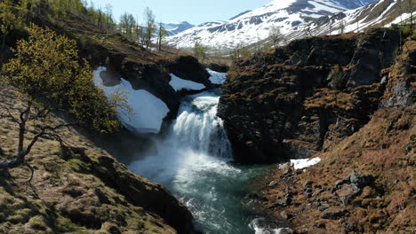 Aerial-view-away-from-a-waterfall-with-snowy-mountain-peaks-in-the-background,-sunny-summer-day,-in-the-Lyngen-alps,-North-Norway---Pull-back,-drone-shot