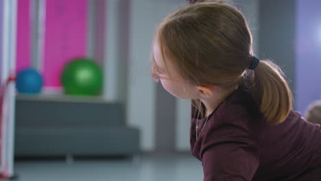 close-up of a young girl with hair tied back, performing an arm stretch exercise, with two exercise balls visible in the background and another child also stretching