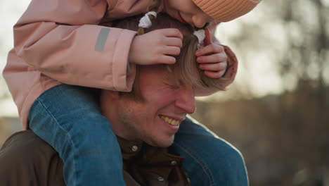 joyful father and daughter moment as the little girl in a pink cap and jacket plays with his hair while sitting on his shoulders