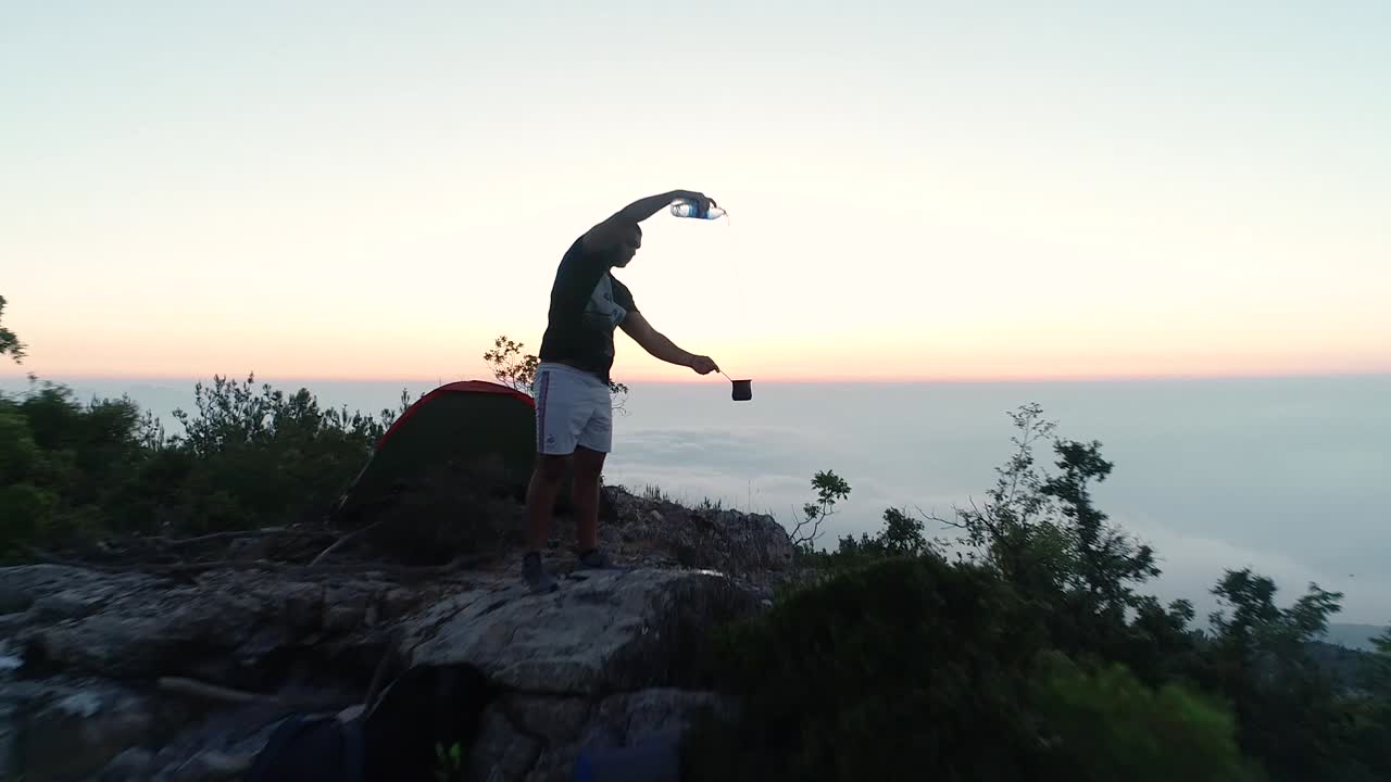 Il drone aereo si avvicina a un uomo sulla cima di una montagna, riprendendo l'acqua che viene versata in una tazza, al rallentatore.