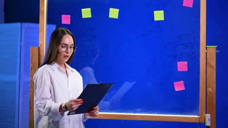 caucasian lady in glasses looks at the clipboard in her hands. glass wall with sticker notes and written numbers behind the girl.