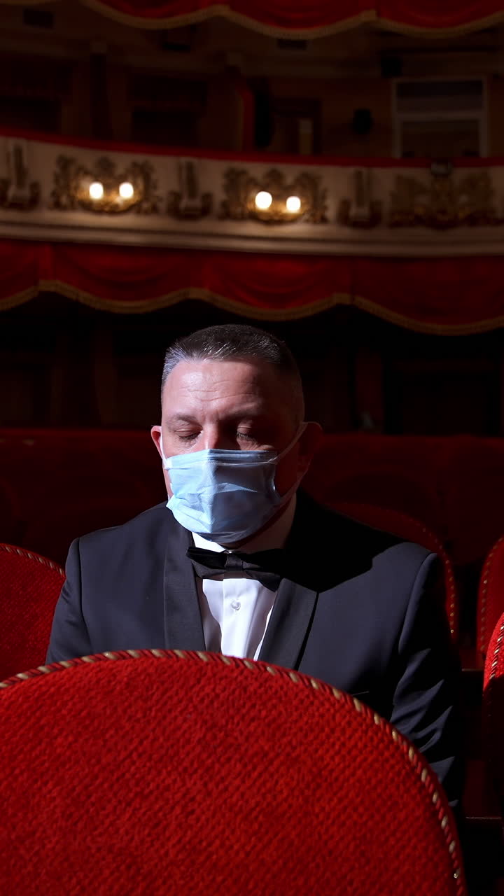 Visitor in suit and medical mask sitting in comfortable chairs alone on dark theater background. Male among rows of empty red chairs. Pandemic concept. Vertical video