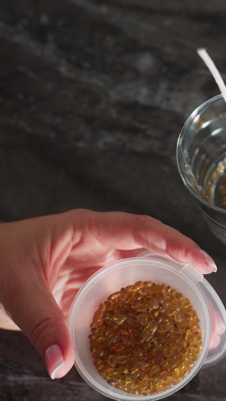 Close up of hand holding small plastic container filled with amber colored gel capsules. background features dark countertop and another glass bowl suggesting preparation or handling