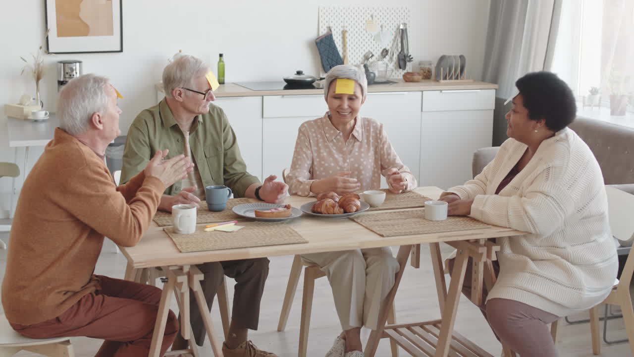 Medium long of diverse multiethnic senior friends with yellow sticky notes on foreheads sitting at dining table, smiling, talking, Asian woman winning, all clapping hands