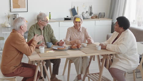 medium long of diverse multiethnic senior friends with yellow sticky notes on foreheads sitting at dining table, smiling, talking, asian woman winning, all clapping hands