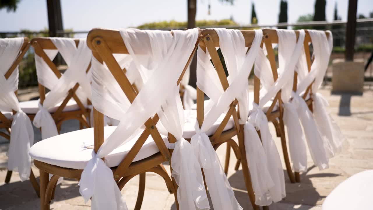 Wooden chairs are elegantly adorned with white fabric at a charming outdoor wedding venue. The sunny atmosphere creates a lovely backdrop for the celebration