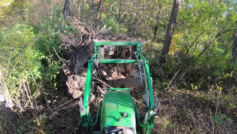 High-angle-point-of-view-on-small-green-tractor-while-using-lift-forks-to-push-tree-stumps-into-a-pile-for-later-burning-in-the-woods-in-autumn