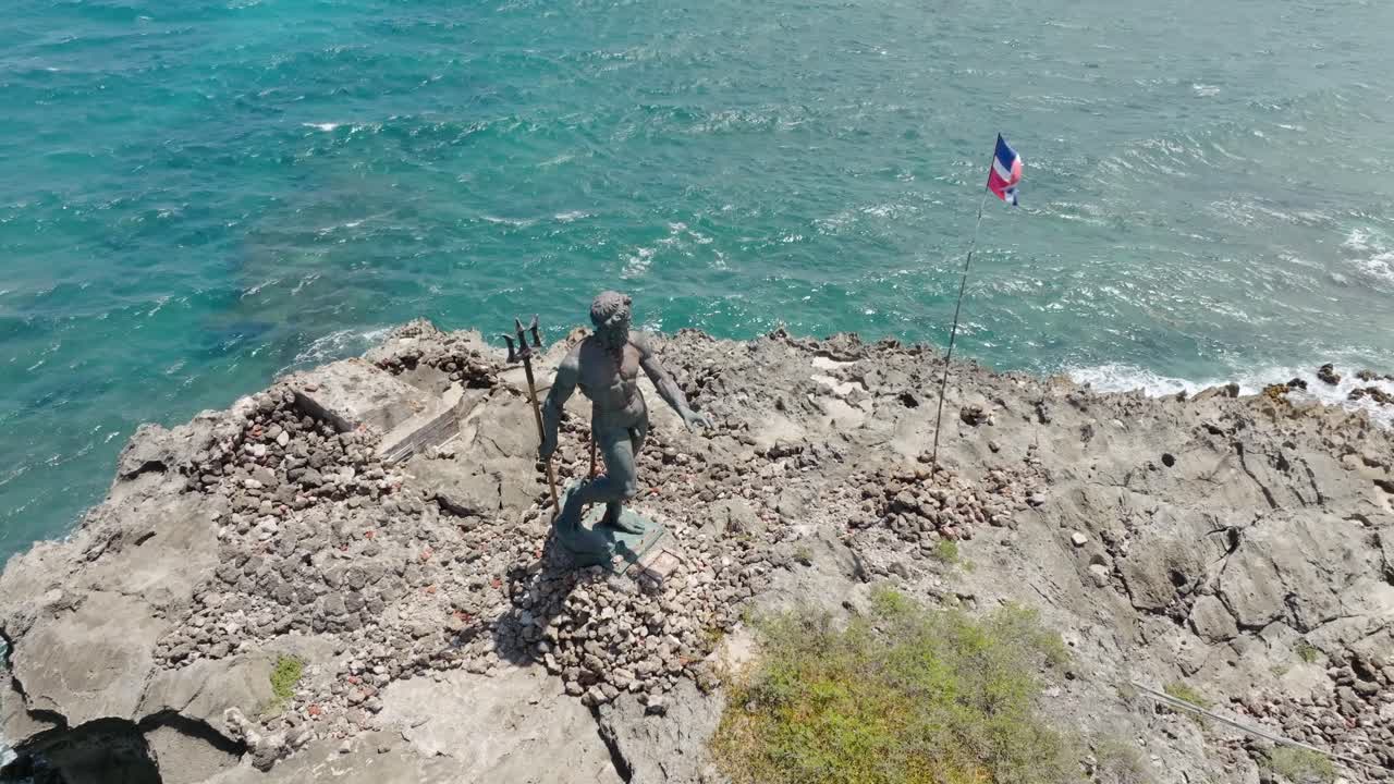 Neptune statue on rocky cliff overlooking sea, bronze sculpture of sea god with Dominican flag, Puerto Plata, Dominican Republic. Aerial tilt-down ascending