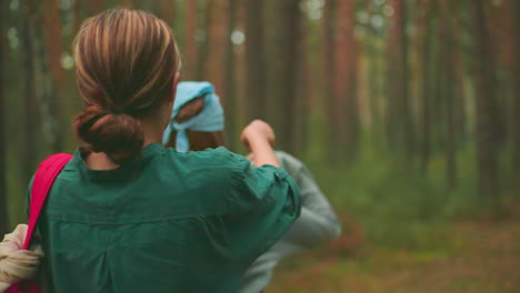 back view of young woman in green shirt helping friend with blue bandana to put on backpack in peaceful forest, sunlight filters warmly through trees