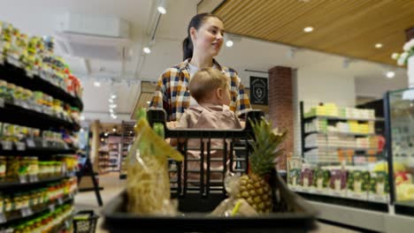 a confident brunette girl in a plaid shirt carries a cart with groceries in which her little daughter is sitting during family shopping in a supermarket