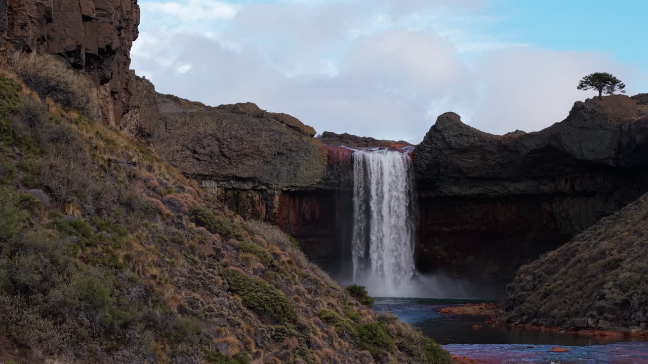 A stunning aerial dolly shot moves from behind a hill to reveal the majestic Salto del Agrio waterfall as it plunges over mineral-stained cliffs in the Patagonian landscape of Argentina