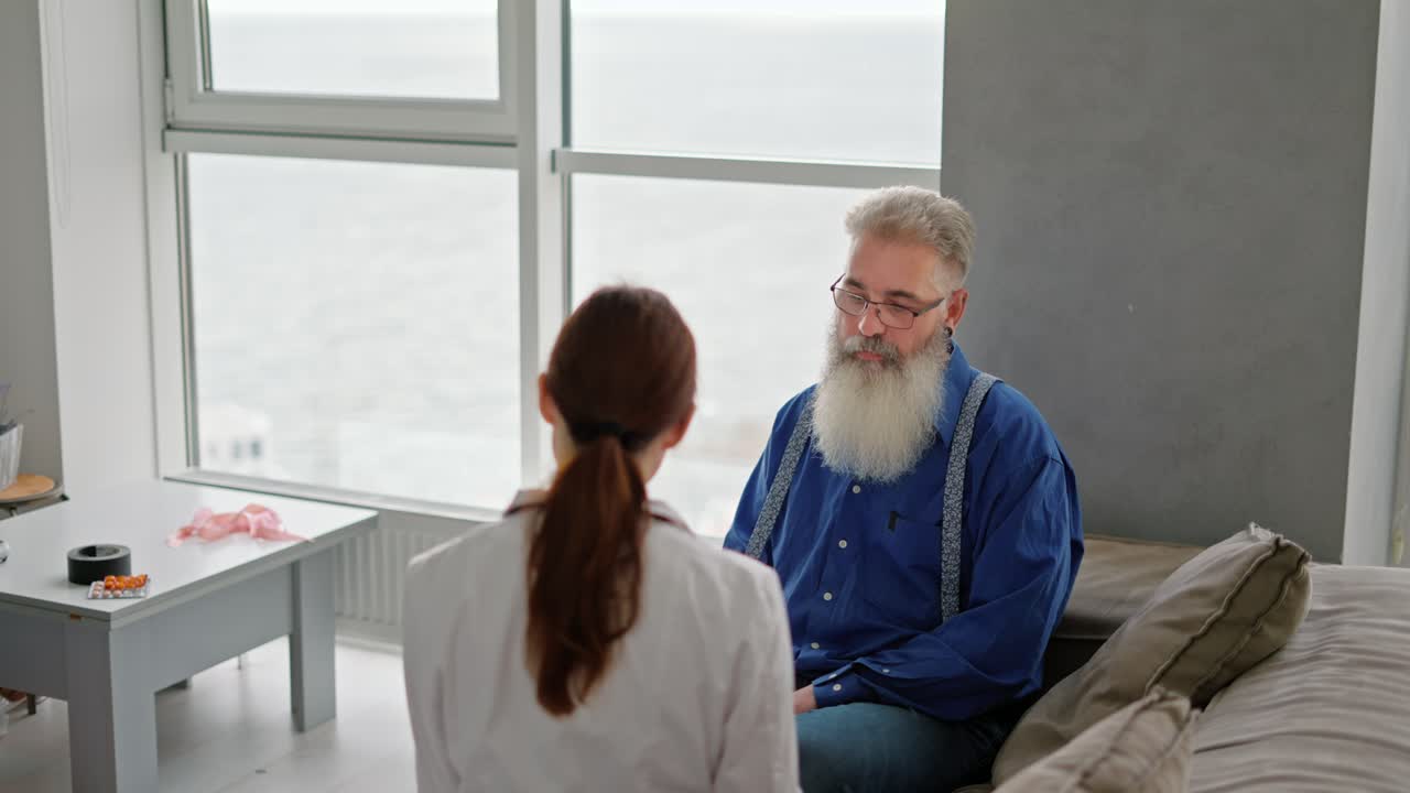 An elderly man with gray hair and a lush thick beard in a blue shirt communicates with a brunette medic girl in a medical gown during a home examination in a modern apartment