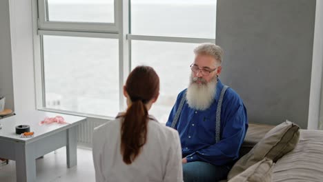 An-elderly-man-with-gray-hair-and-a-lush-thick-beard-in-a-blue-shirt-communicates-with-a-brunette-medic-girl-in-a-medical-gown-during-a-home-examination-in-a-modern-apartment