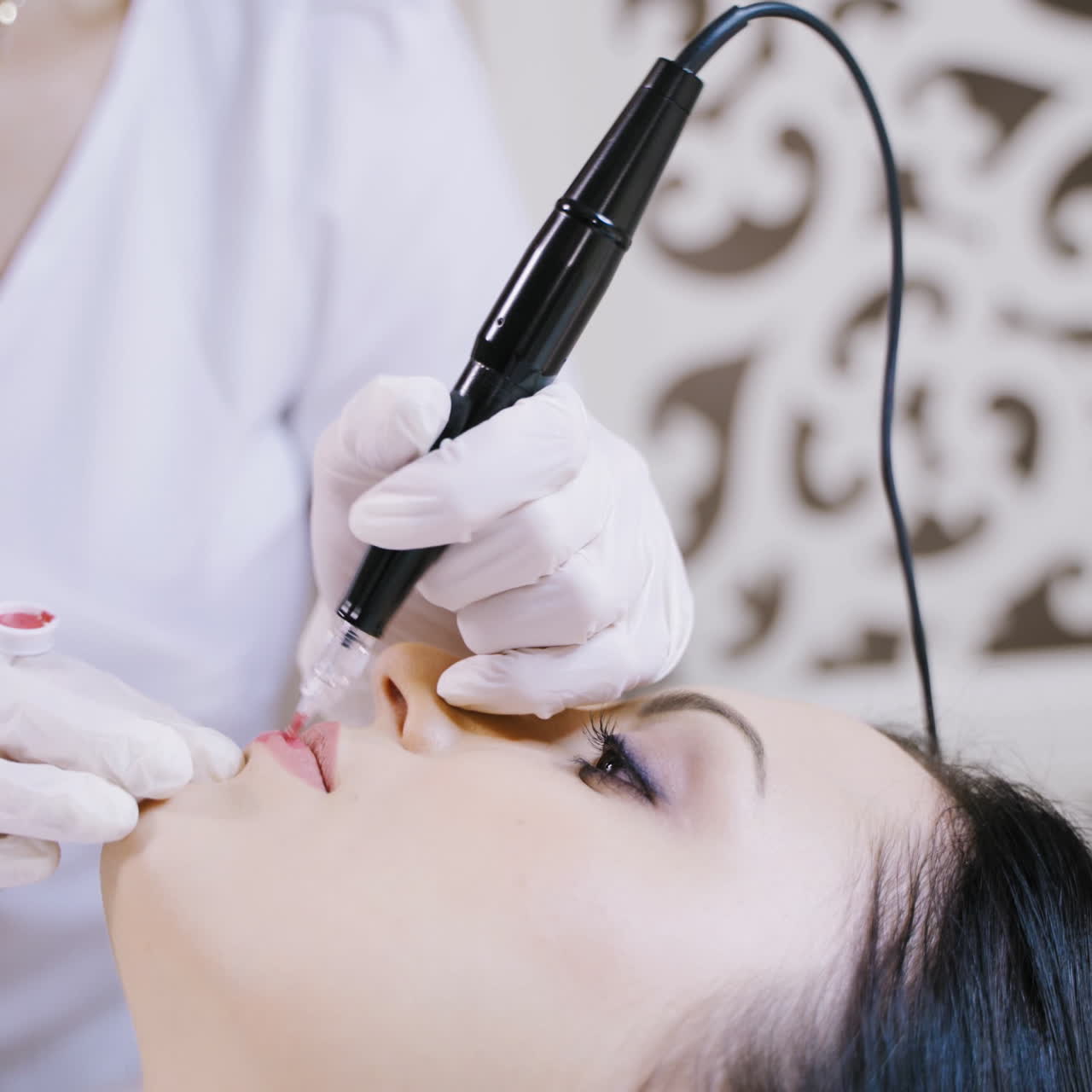 Skilled cosmetician applying permanent make-up on lips in a beauty salon. Young woman undergoing procedure of permanent lip makeup with tattooer machine. Close-up