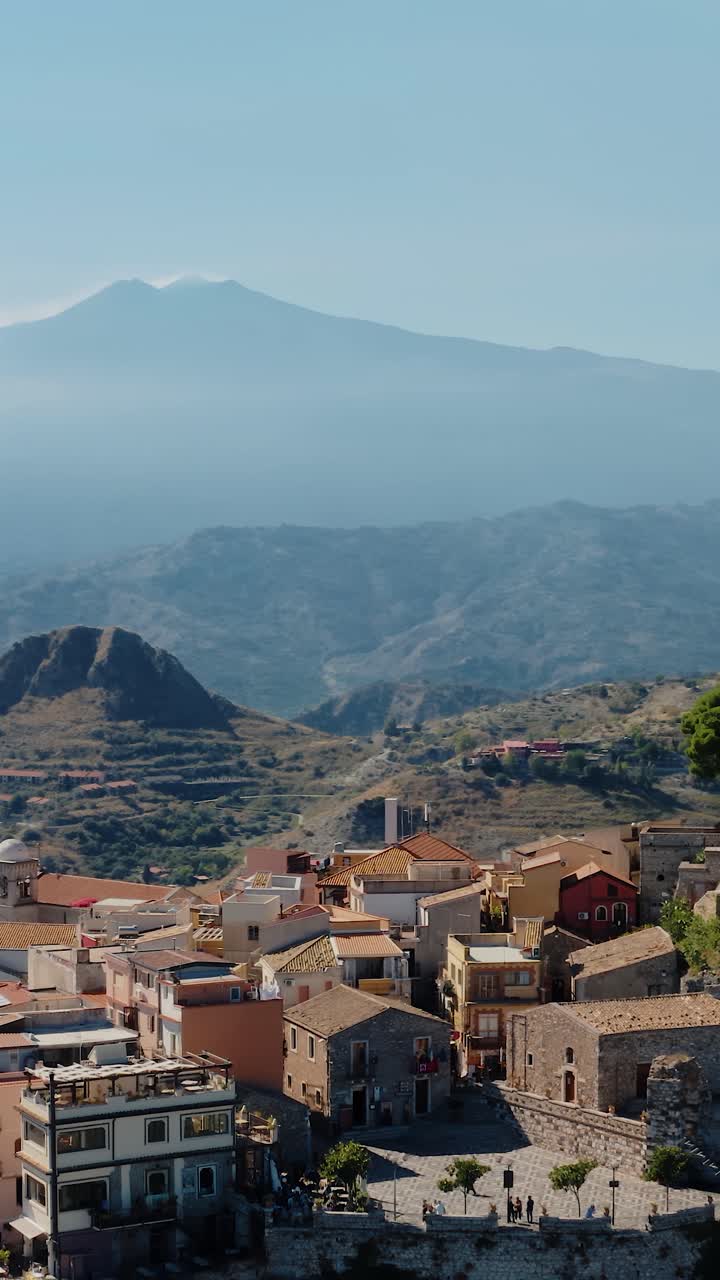вертикальный орбитальный дрон, застреленный над историческим городом кастельмола, построенным на естественной террасе на краю скалы с горой этна на расстоянии. итальянская деревня в горах. дым, исходящий из вулкана.