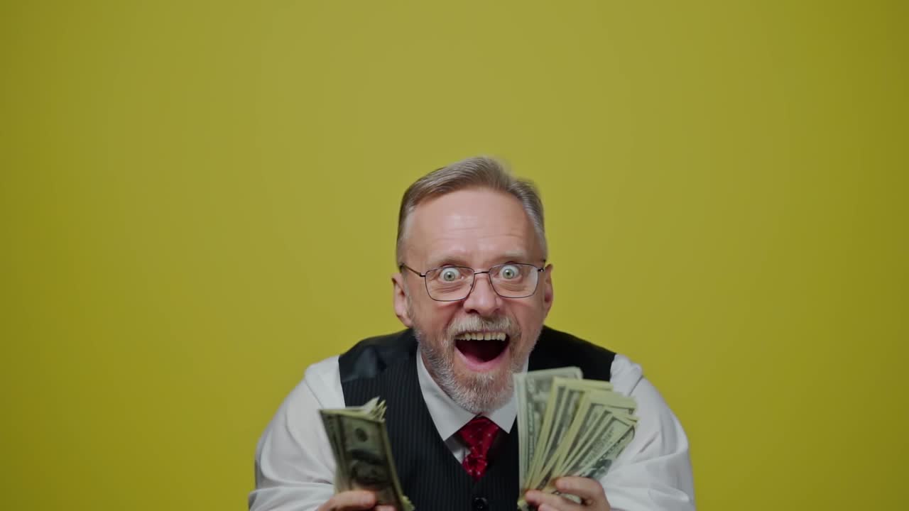 Joyful old man holding banknotes in his hands. Funny senior man with widely open eyes and mouth holds dollar bills on the yellow background. Positive emotions.