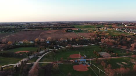 aerial drone flying over rural countryside in wisconsin with the view of agricultural fields, baseball sports field during a sunny summer evening time