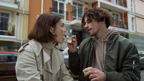 Lovely-couple-spending-date-on-street.-Man-feeding-woman-on-city-background.