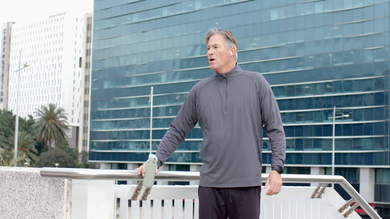 Senior man recovering from run on rooftop, placing bottle on railing and doing quadriceps stretch