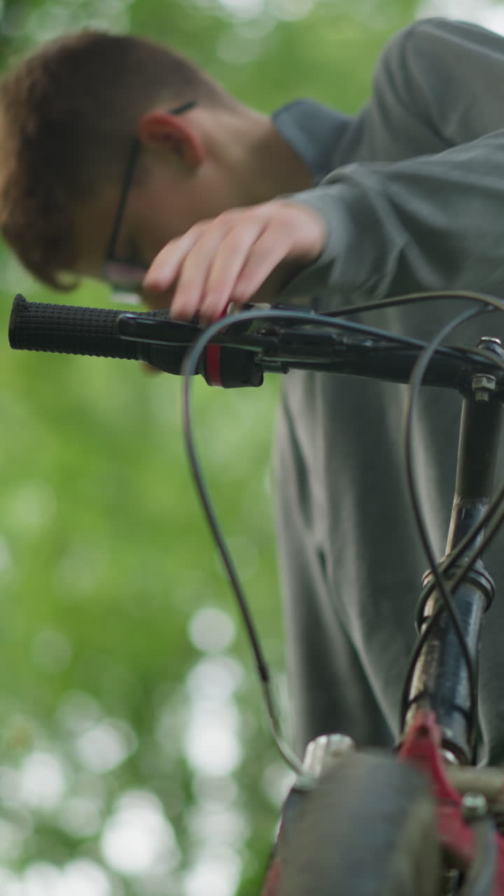 niño con gafas sostiene el manillar de una bicicleta estacionada en un campo cubierto de hierba, agarra el manillar con firmeza, comprobando la funcionalidad de los frenos mientras está de pie, la bicicleta está estacionada cerca de un árbol