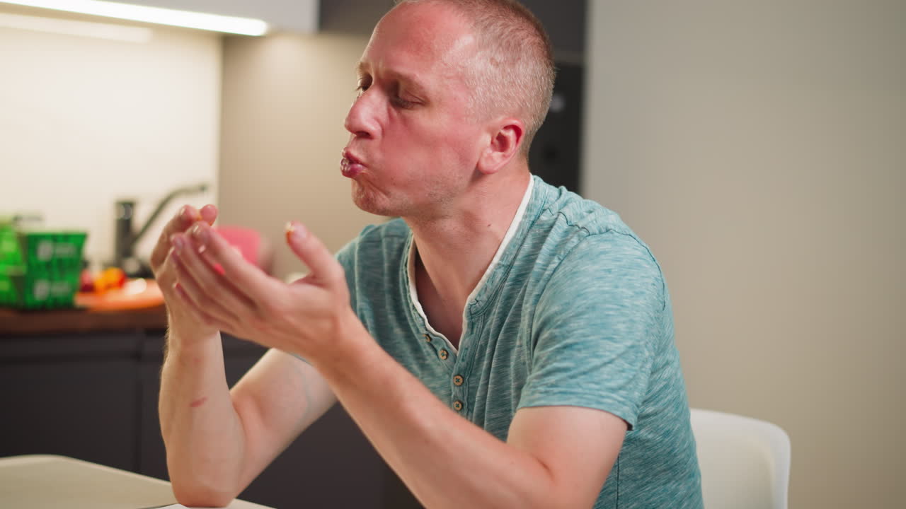 White man wearing teal shirt finishes eating, licking mouth and fingers at table with blur kitchen background, savoring last taste, casual dining moment filled with satisfaction and enjoyment