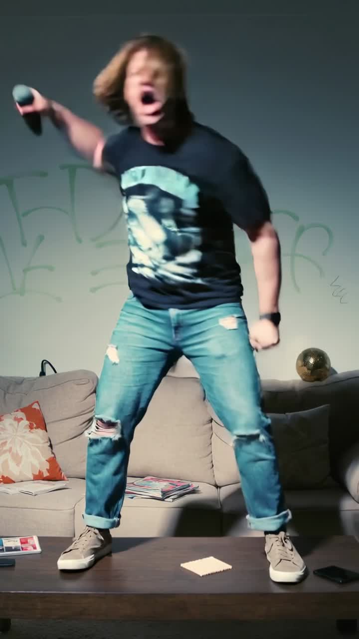 Video verticale: Telecamera che riprende un uomo che fa headbanging e fa oscillare un microfono su un tavolino da caffè a casa, con una maglietta di una band.