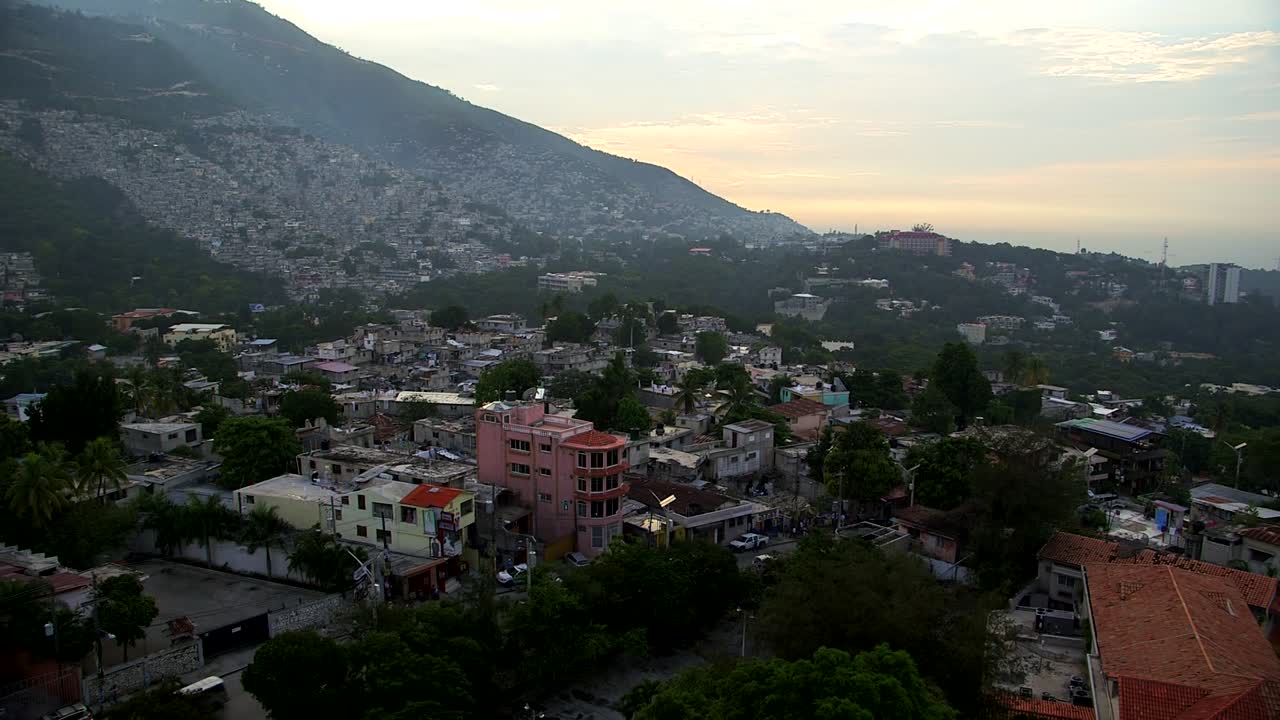 Utsikt over åssidene i Port-au-Prince på Haiti ved solnedgang, med tette hus som klatrer oppover fjellet, spredte trær og disig karibisk himmel over bukten