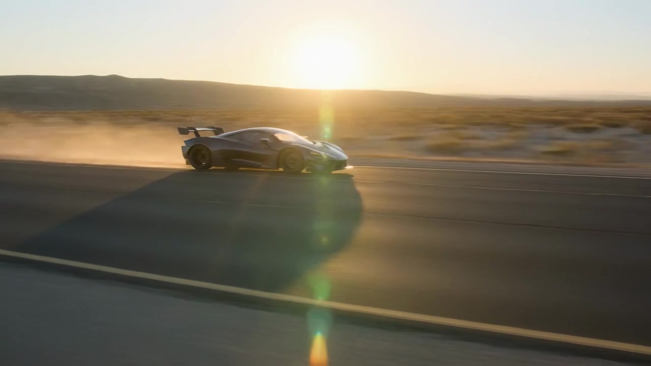 Entrando da sinistra, un'auto sportiva veloce corre lungo un'autostrada nel deserto, sollevando polvere, mostrando l'alettone posteriore.