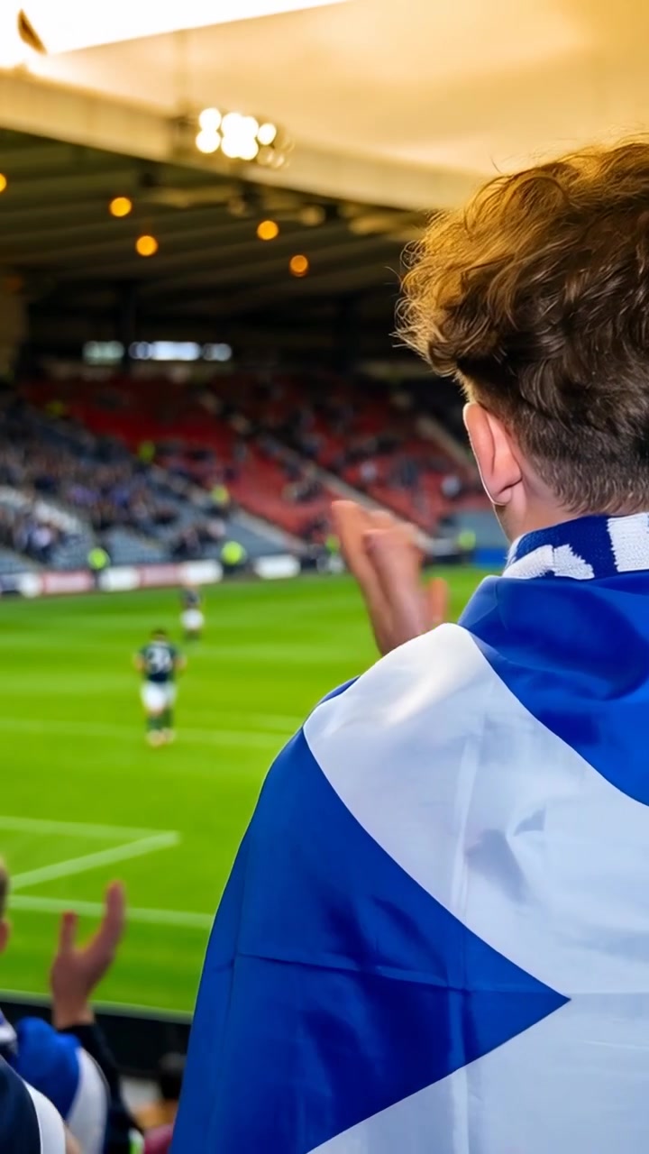 Video verticale: Tifoso avvolto nella Saltire che guarda il campo mentre un amico applaude durante il gioco, con spazio per il testo.